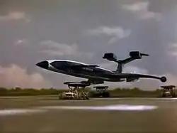 A gleaming, blue, futuristic airliner rests on top of three orange support vehicles (one underneath the fuselage and two underneath the wings) as it speeds out of control along a runway against a grassy backdrop, the sky bright with clouds.