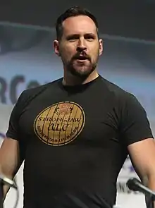 A black-haired man wearing a black shirt with a single yellow circle.