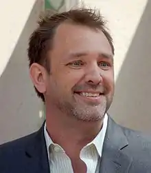 A brown-haired man wearing a lime shirt and grown jacket smiles and looks at something off-screen.