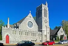 Two pointed gray stone buildings seen from across a street, with cars passing in front and a traffic light visible from the center to the upper left. The one on the left has a tall square stone tower with a smaller tower topped by a cross at the top. The one on the left has a large round window in the middle and a cross at the point on the front. Both have open red doors.