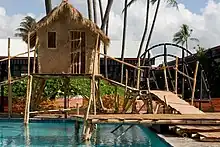 A grass and bamboo hut is sitting above a swimming pool due to bamboo stilts. A bridge made of wood is leading up from the right towards the hut. In the background is other buildings and blue, cloudy skies.