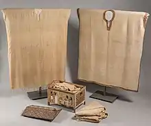 Two tunics on a stand with other items folded inside a chest and piled on the floor beside it