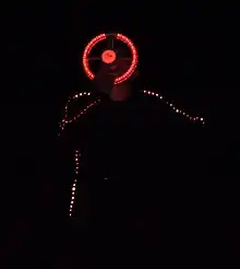 Close up, the dark-clothed singing man has lights embedded in his jacket sleeves and side, and is singing into a microphone with an illuminated circular structure around it, somewhat like a steering wheel, that he is holding with one hand.