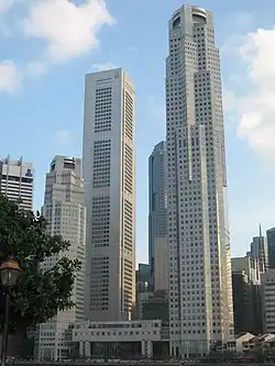 Ground-level view of several skyscrapers; the building on the right is the tallest and has a square cross section with several diagonal protrusions. A shorter building on the left resembles it closely.