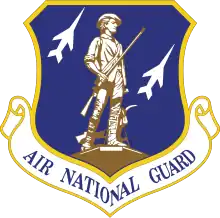 A blue shield with yellow outlining emblazoned with a man in 18th-century clothing holding a rifle in brown. Behind him are two profiles of fighters in white. Below the shield is a white scroll with yellow outlining that states "Air National Guard".