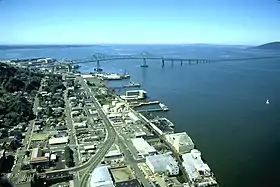 A long bridge crosses a huge river flowing into a vast body of water under a blue sky. The bridge begins in a settlement with streets, buildings, and wharfs along the river and extends out of sight toward a low hill on the far shore. The first part of the bridge has a superstructure and is high above the water but then the bridge gradually descends and continues out of sight not so far above the water.