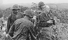 A black-and-white image of Merritt Edson and three other Marines standing in bushes. Edson is holding some binoculars and is looking out over the battlefield.