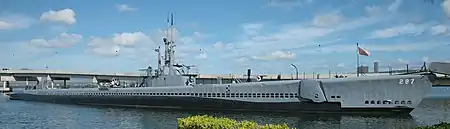 A submarine, long, sleek and painted grey at anchor. It flies a red and white striped ensign.