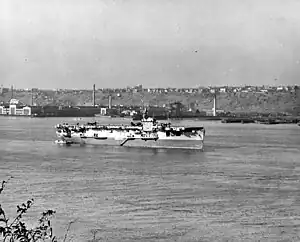 A World War II-era aircraft carrier at anchor. Several aircraft are on her flight deck and land and buildings are visible in the background.