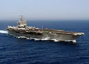 A large gray ship with a flat surface and tower in the middle traveling through the sea