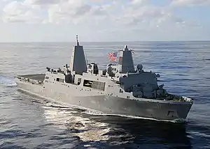 A grey ship sailing on the blue ocean, flying an American flag from its mast. Behind it are clear and sunny skies.