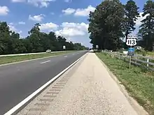 A four-lane road with a grassy median surrounded by farms and woods