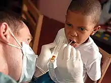 Man holding plastic tray with brown material in it and sticking a small stick into a boy's open mouth