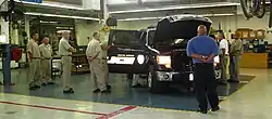 A utility vehicle in a workshop surrounded by men, most of whom are wearing identical beige uniforms