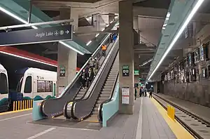 Inside an underground train station at platform level with a set of escalators leading upward and a train in the background.