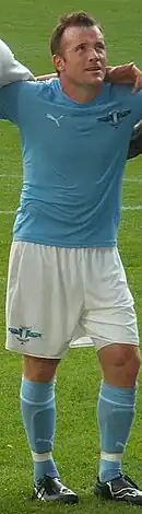 A dark haired man stands with his arms around the shoulders of two teammates, each of whom is out of shot. The subject wears a light blue shirt, white shorts and light blue socks.