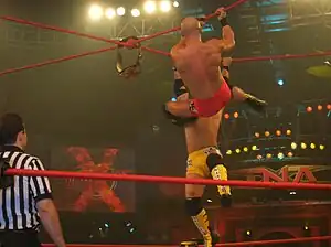 Two men, one wearing red trunks and the other wearing yellow trunks, hanging from red crossed ropes that form an "x" above a wrestling ring as a referee watches.