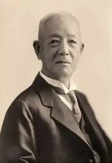 Black and white portrait. Head shot of Iwadare in front of a light background, wearing a dark suit jacket with a light wing-collared shirt, dark vest, and dark polka-dotted necktie.