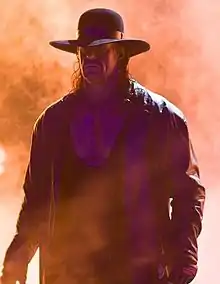 A man with long dark hair, wearing a black wide-brimmed hat, black leather jacket, and black wrestling tights.