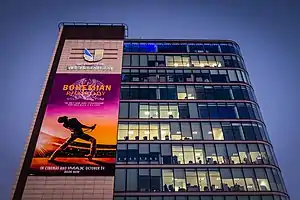 A building in London with its electronic wall displaying the poster for Bohemian Rhapsody. Around it are blue skies.