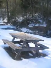 A wooden picnic table in the snow, with a small stream and bare and conifer trees in the background