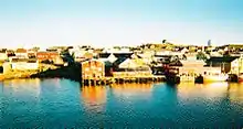 A cluster of buildings and wharves, with a small ship moored to the right of picture. The waterside buildings are clearly reflected in the sea.