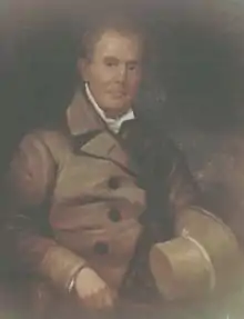 portrait of McBee while seated, wearing a brown coat with dark buttons over a white shirt collar against a dark background, and holding a matching hat on its side over his lap with his right arm resting across the furniture arm