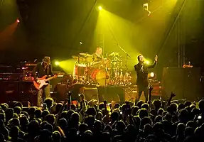 Three white Englishmen perform on a darkened stage, with yellow lights shining on them, whilst a crowd watches them.