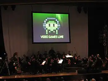 A photograph of an orchestra on a dimly lit stage. Above the group is a projection screen with a black, white, and green image of pixel art. The pixel art is an oval object wearing headphones with eyes and four tentacles. Below the pixel art is the phrase "Video Games Live".