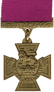 A medal in the shape of a cross hanging from a crimson red ribbon.