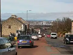 A busy main road with a distant view of an urban landscape. On the left are two-storey buildings; on the right are some mature trees.