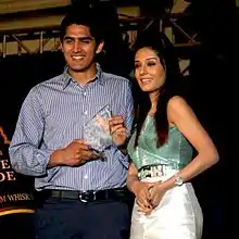 An young Indian male and a female standing side-by-side. The man is on the right and wears a blue-grey shirt and navy-blue trousers. He is smiling looking down to the right of the camera and holds a glass, triangular shaped trophy in his hands.  The female also looks towards the bottom right and wears a shiny dress whose top portion is light green in color and the skirt area being silvery. Her long black hair falls on her shoulders.