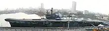 A small aircraft carrier tied up at a wharf. A helicopter and two aircraft sit on the flight deck. Other ships and skyscrapers are in the background.