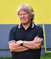 Middle-aged white man wearing dark polo shirt, standing with arms crossed, squinting to the right of camera