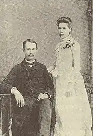 A black-and-white photograph of a couple. On the left is a seated man with a moustache weraing a dark suit. Standing on the right is a woman in a white dress, body facing left.  Both face the camera.