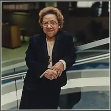 A woman in a dark suit, standing with her arm resting on a glass railing in a library.