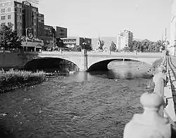 A concrete arch bridge crosses a river in two arch spans, with mid-size buildings visible in the distance.