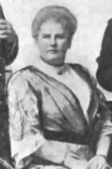 A white woman with light-colored bouffant hair, seated, wearing a loose satiny robe-like dress over a white high-collared blouse