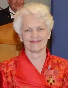 Head and shoulders portrait of a smiling white-haired woman in a red dress, with an order pinned to her lapel.