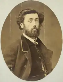 Chest-high portrait of man in his twenties with brown eyes and dark medium-long hair and a beard, wearing a vest and suitcoat and white shirt