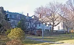 A large light house with a peaked roof, wings and many dormer windows seen from down a slope. There is a large bare tree in front of its balustraded deck on the right