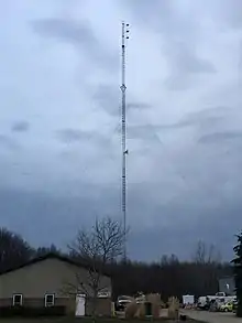 An alternating white and red triangular guyed lattice tower, set against a cloudy sky. A three-bayed antenna at the top of the structure is also flanked by an activated red beacon light. An additional red beacon light is fixed on the structure above a small studio-to-transmitter link dish. An office building is pictured in front of the tower on the left-hand side, with a parking lot on the other side.