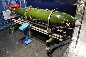 A mostly green coloured bomb around 3 metres long, marked with the word "training" and with 4 fins at the rear