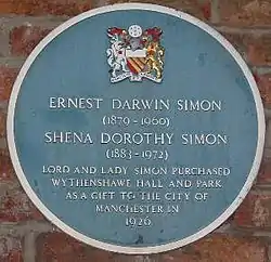 A circular plaque coloured blue mounted on a brick wall, containing the coat of arms of Manchester City Council at the top, the white text "Ernest Darwin Simon (1879–1960)" and "Shena Dorothy Simon (1883–1972)" in the centre, followed by "Lord and Lady Simon purchased Wyhtenshawe Hall and Park as a gift to the City of Manchester in 1926"