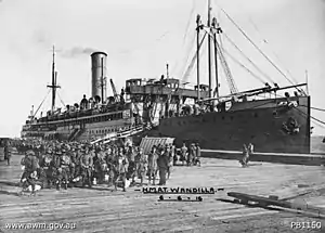 A crowd of diggers in slouch hats pose in front of a ship docked at the wharf.