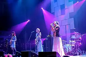 Four female musicians performing live on-stage against a black and white backdrop with violet lights.