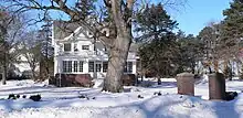 House with carriage barn in background. Large deciduous tree in front of house; square brick piers in foreground