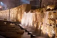 To the right of some stepping stones, a row of lamps illuminates a short artificial waterfall streaming down a honey-colored wall made of limestone bricks.