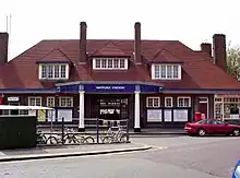 A brown-bricked building with a red-tiled roof and a rectangular, dark blue sign reading "WATFORD STATION" in white letters all under a white sky