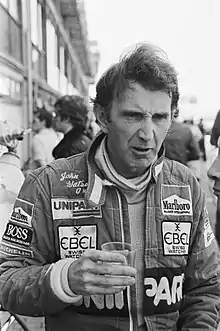 John Watson in racing uniform holding a glass of water in his right hand